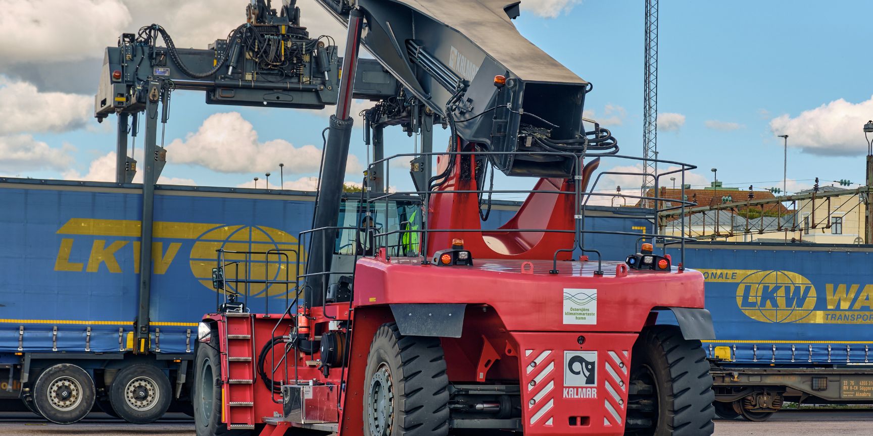 Nahaufnahme eines massiven Krans in einem DUSS-Terminal, der einen blauen LKW-Sattelauflieger präzise anhebt. Der Greifarm hält den Auflieger in der Luft, bereit zur Verladung auf einen Güterzug. Dies visualisiert den Umschlagprozess Schiene-Straße im Kombinierten Verkehr.
